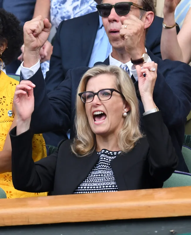 A woman in glasses and a black patterned dress enthusiastically cheers with others at an event.