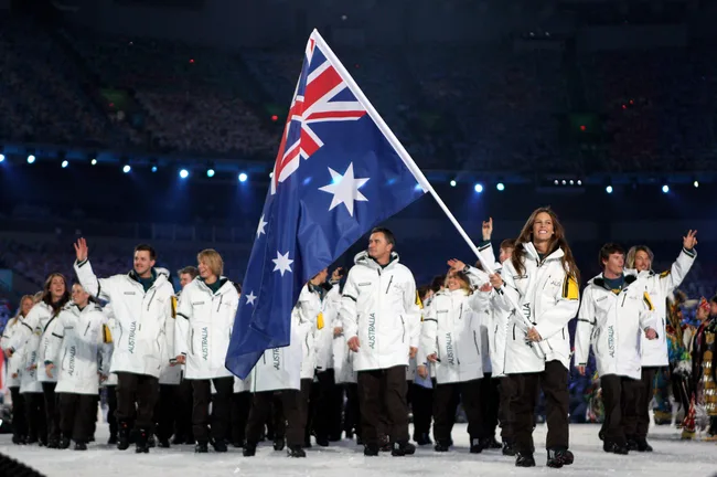 Australian athletes marching with flag at Winter Olympics opening ceremony, wearing white coats, smiling.