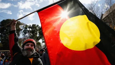 Man holding Aboriginal flag at outdoor event, sunlight shining through flag.