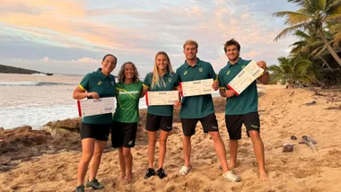 Five athletes holding tickets pose on a sandy beach at sunset, with palm trees and ocean in the background.