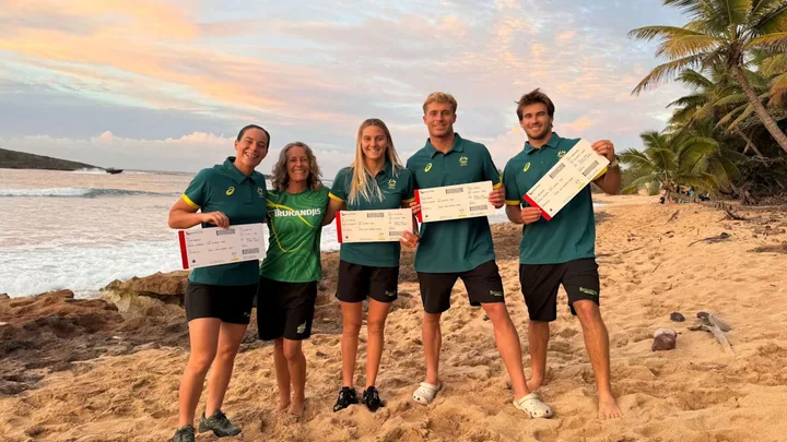 Five athletes holding tickets pose on a sandy beach at sunset, with palm trees and ocean in the background.