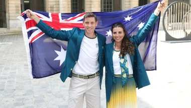 Australian athletes pose with flag, wearing green blazers and smiling, outside on a cobblestone street.