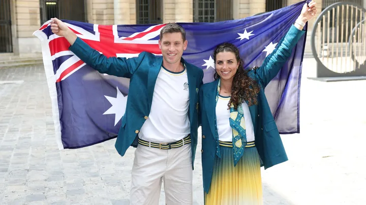 Australian athletes pose with flag, wearing green blazers and smiling, outside on a cobblestone street.