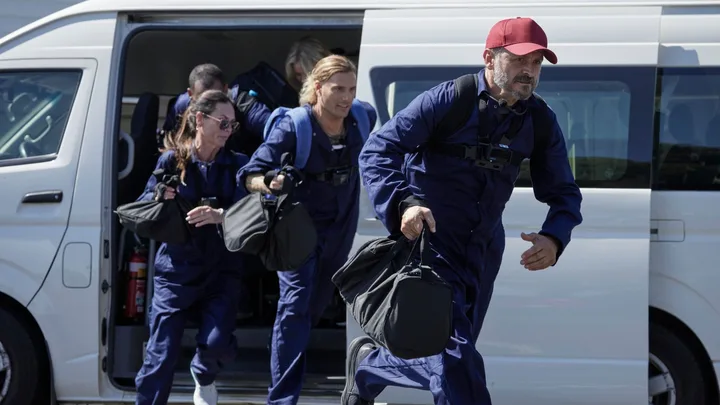 Group in blue uniforms, exiting a van with bags, one person leading with a red cap.