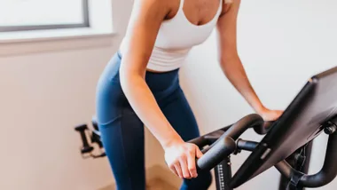 Person in workout clothes using a stationary exercise bike indoors, holding handlebars and leaning forward.