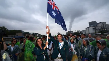 Australian team members in rain ponchos hold the national flag during a parade on a cloudy day.