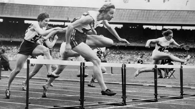 Athletes competing in a hurdle race at a large stadium, with spectators in the background.