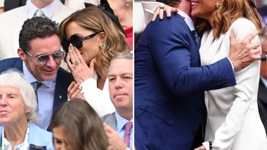 Two people in elegant attire, whispering and embracing warmly at a crowded event.