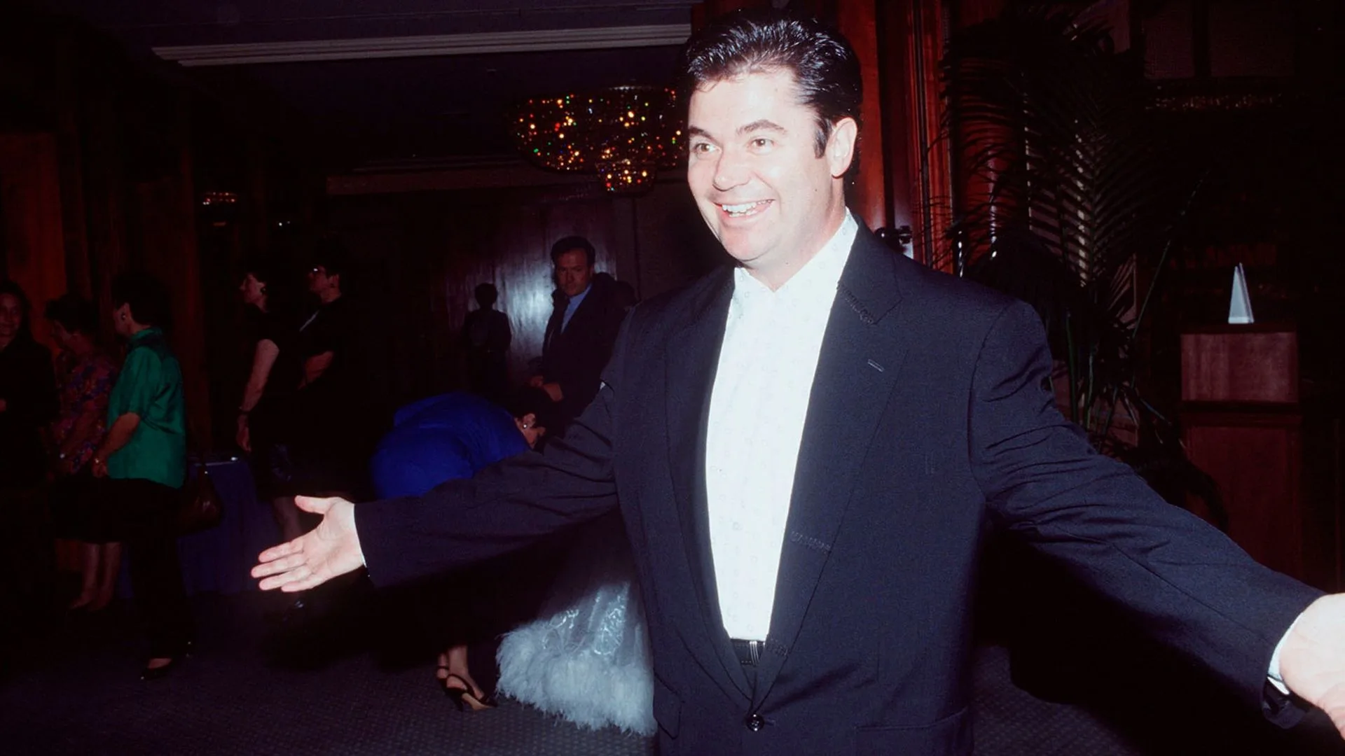 A man in a black suit smiling with arms open in a dimly lit room with people in the background.