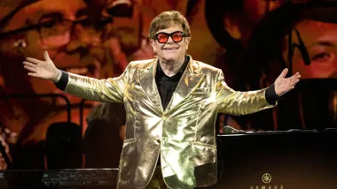 A person in a golden suit with red glasses performs on stage, smiling with arms open wide.