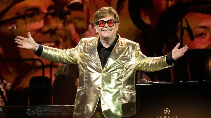 A person in a golden suit with red glasses performs on stage, smiling with arms open wide.