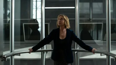 A woman with short blonde hair stands in a glass elevator, looking up, with her hands on the handrail.