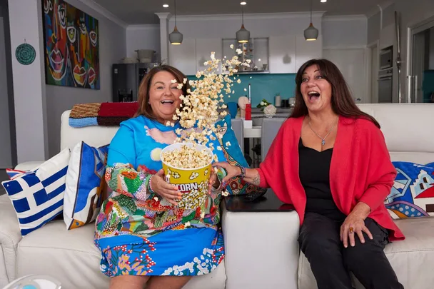 Two women laughing on a couch, spilling a bucket of popcorn enthusiastically in a colorful room.