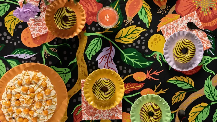 Colorful table with donuts, cake topped with berries, vibrant floral-patterned cloth, napkins, and a candle.