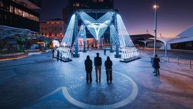 A group of people viewing a large geometric light installation in a city square at night.