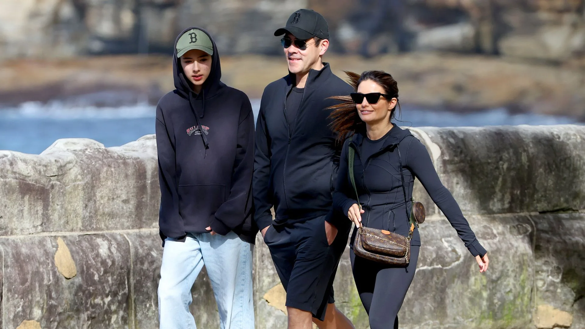 Group of three people walking outdoors on a sunny day, dressed casually in hoodies and sunglasses.