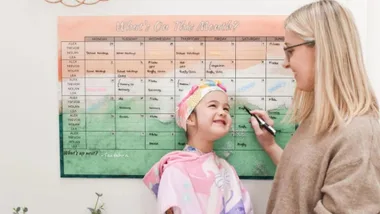 A woman writes on a monthly planner as a smiling child looks on, wearing a colorful towel wrap.