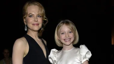 A woman in a black dress and a smiling young girl in a white dress pose together against a dark background.