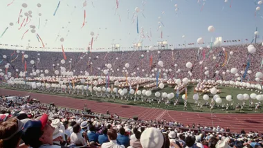 A stadium filled with spectators and performers, balloons and streamers fill the sky, celebrating an event.