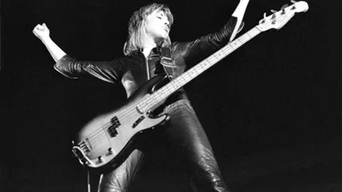 Musician playing a bass guitar on stage, wearing leather outfit, arms raised in a powerful pose against a dark background.