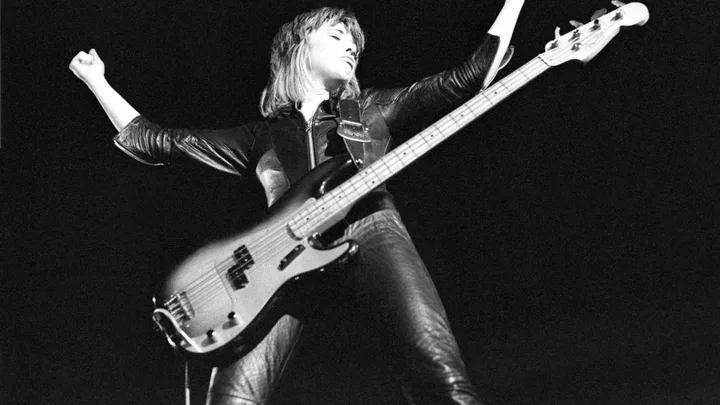 Musician playing a bass guitar on stage, wearing leather outfit, arms raised in a powerful pose against a dark background.