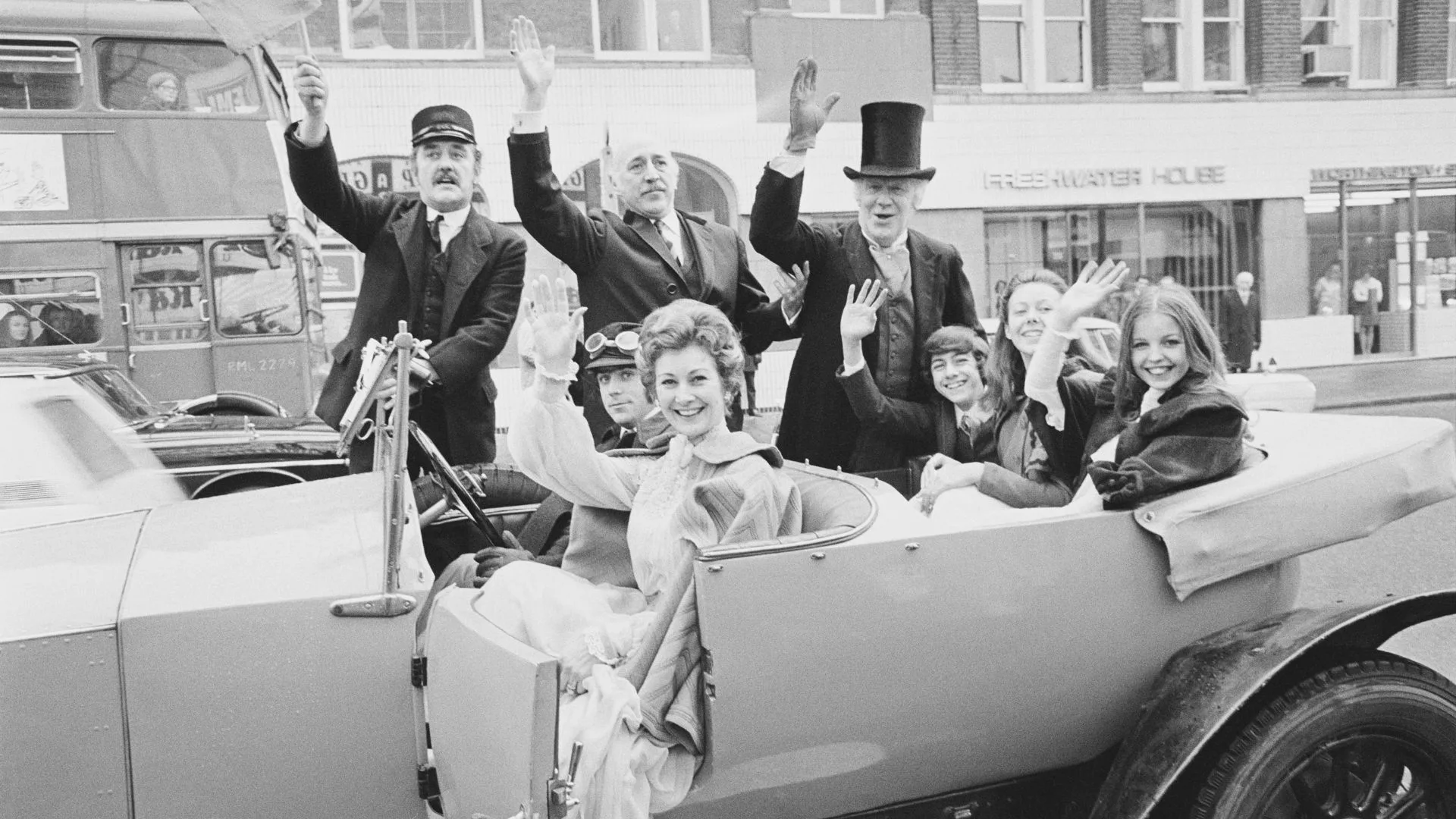 Black and white photo of people waving from a vintage car, with a double-decker bus in the background.