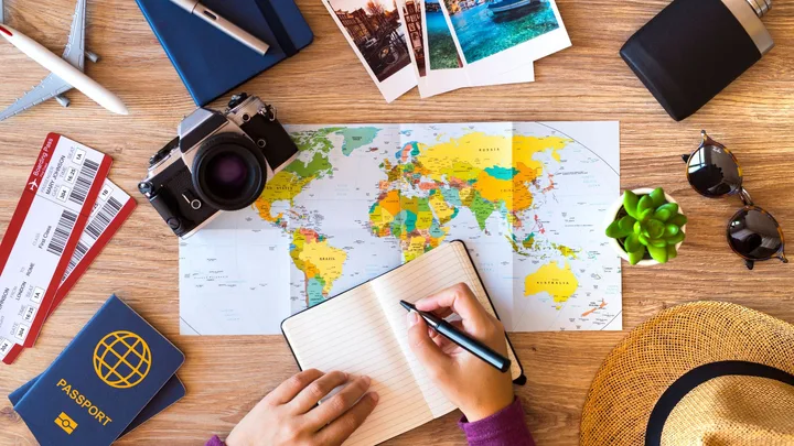 Person planning trip with map, notebook, camera, passport, boarding passes, and travel items on a wooden table.