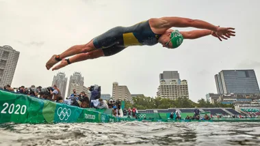 Olympic triathlete dives into the water at the Tokyo 2020 games, buildings and spectators in the background.