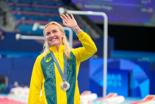 Athlete in yellow and green jacket with a silver medal waves, smiling, indoors at a sporting event.