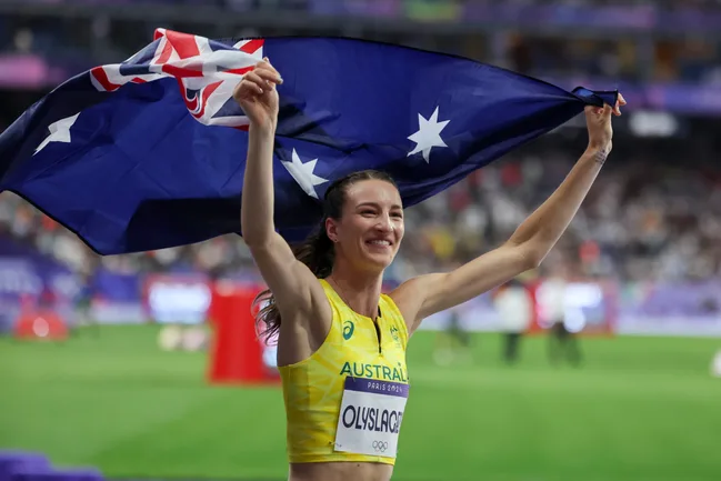 Athlete celebrates with Australian flag in a stadium, wearing a yellow jersey with "Olyslagers" written on it.