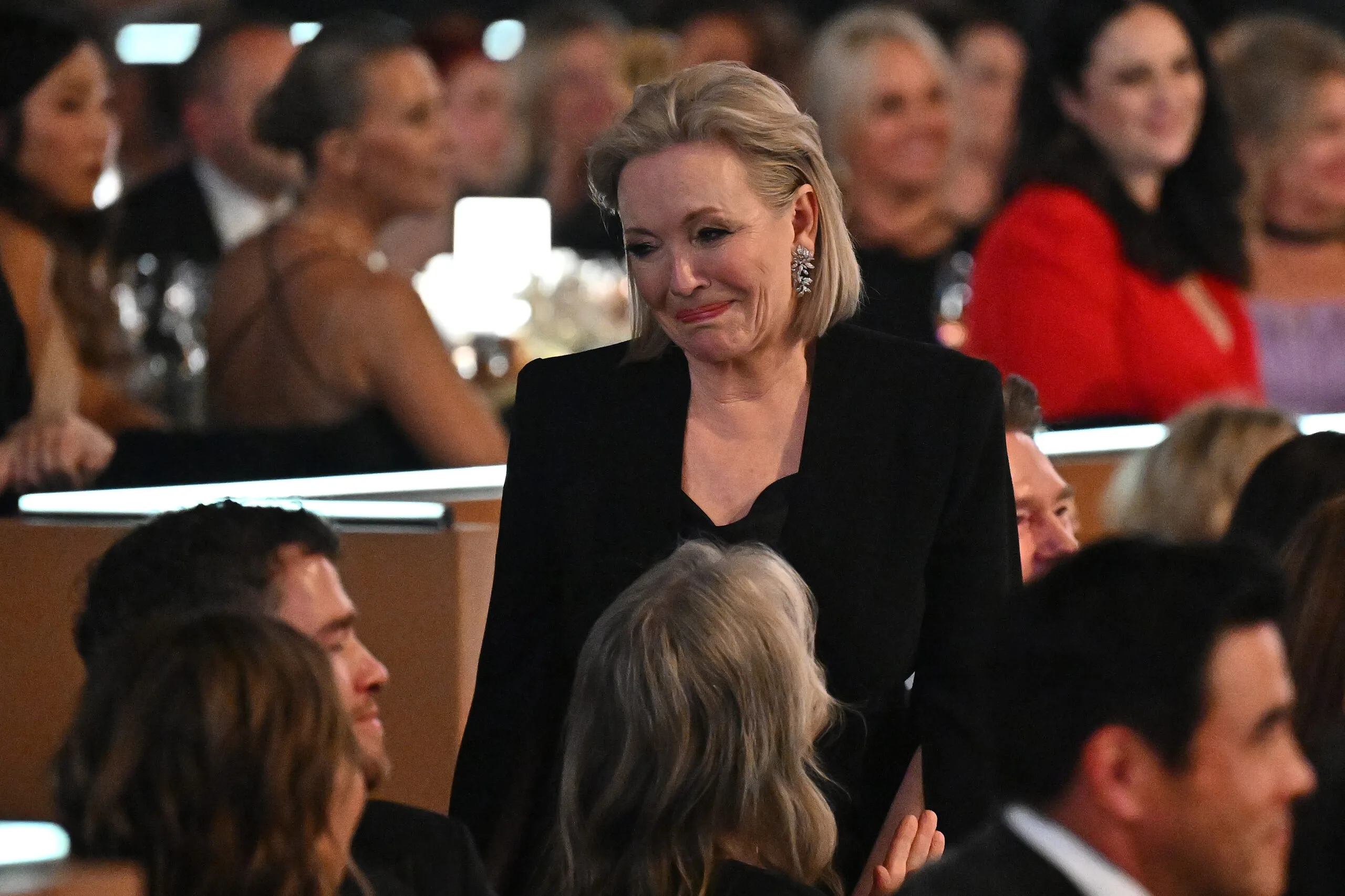 A person in formal attire standing among seated guests at an event, with a warm expression.