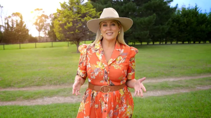 Woman in a floral orange dress and wide-brimmed hat standing on grass, trees in background, sunlight streaming through.