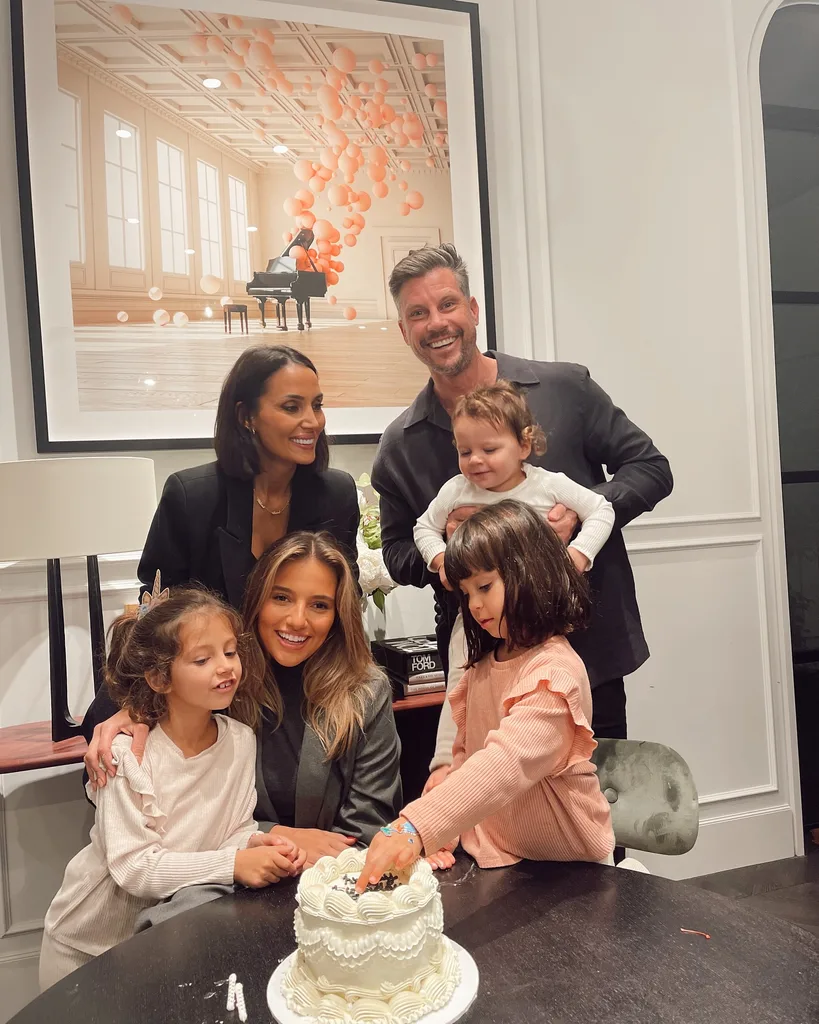 A family is gathered around a table with a white cake, smiling and posing for a photo in a warmly lit room.