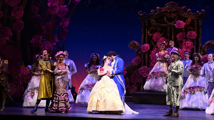 A couple in elaborate costumes share a kiss on stage, surrounded by performers in floral dresses and suits.