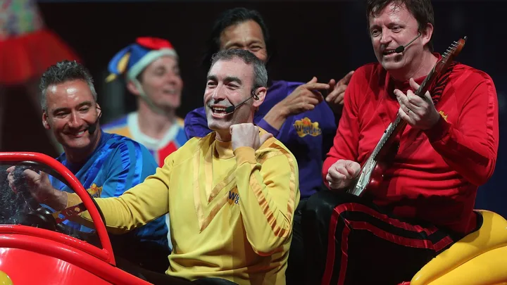 The Wiggles perform on stage in colorful outfits, with one member playing guitar.