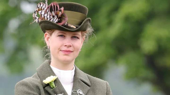 A person in a green outfit and feathered hat, outdoors with blurred greenery in the background.