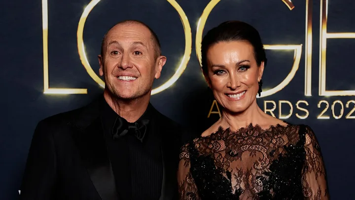 Two individuals smiling at an awards event, one wearing a black suit, the other in a lace dress, against a logo backdrop.