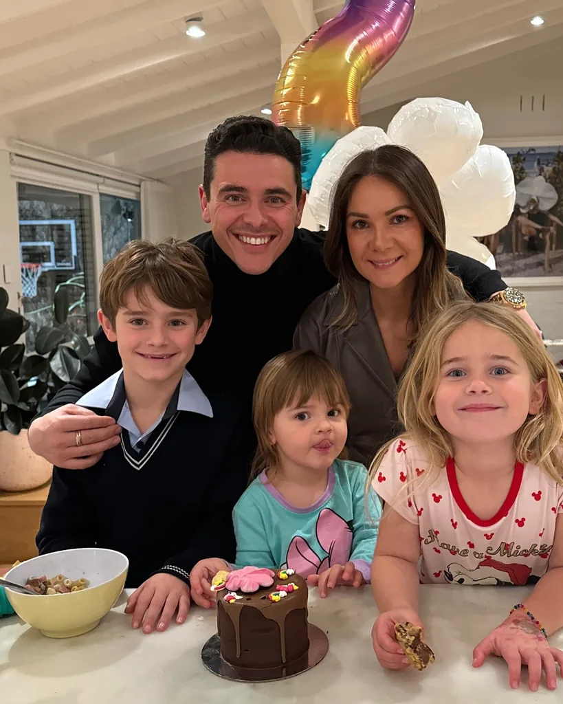 Family of five smiling, gathered around a table with a small chocolate cake and balloons in the background.