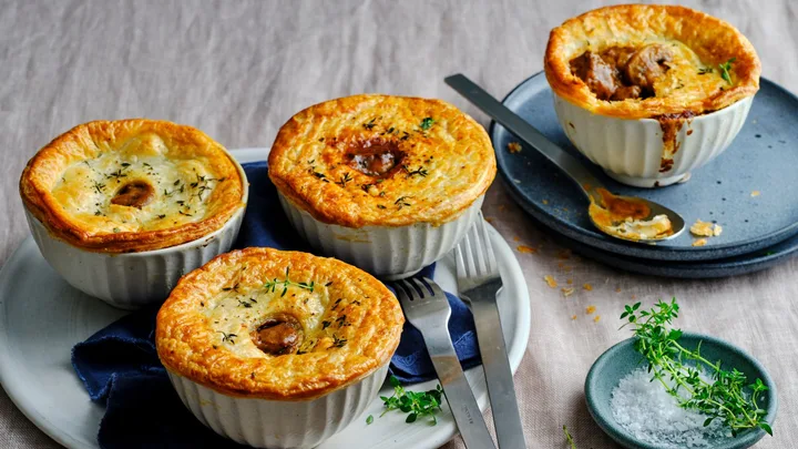 Four savory mushroom pot pies with golden crusts, garnished with thyme, served in white ramekins on a plate with cutlery.