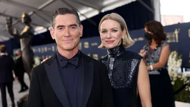 A man and woman in formal attire pose at an event, with a large statue visible in the background.
