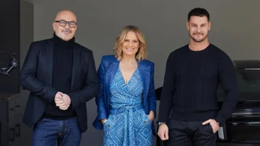 Three people stand smiling, with one man in a black suit, a woman in a blue outfit, and another man in a black sweater.