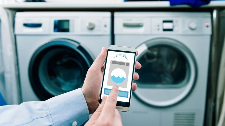 "Person using smartphone to control washing machine with app interface, washers in background."
