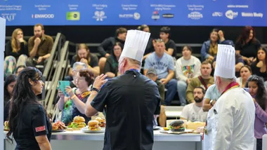 Chefs in white hats presenting burgers to an audience at a cooking show, with a blue sponsor banner in the background.