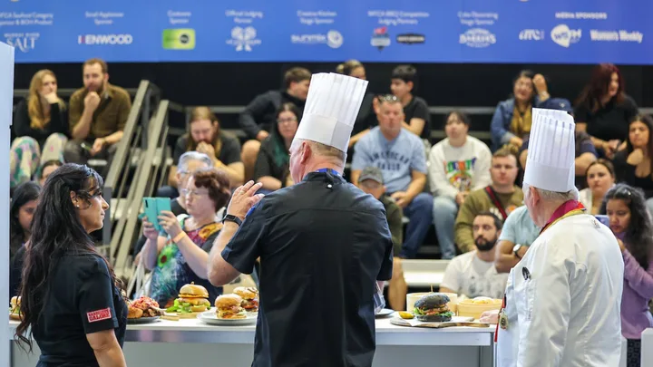 Chefs in white hats presenting burgers to an audience at a cooking show, with a blue sponsor banner in the background.
