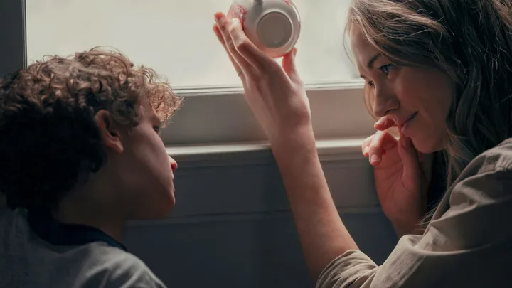 A woman and a boy sit by a window, the woman holds a tea cup with lipstick marks, both appear thoughtful.