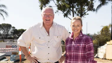 Mature man and woman smiling at a construction site, he wears a light shirt, she wears a plaid shirt; trees in background.