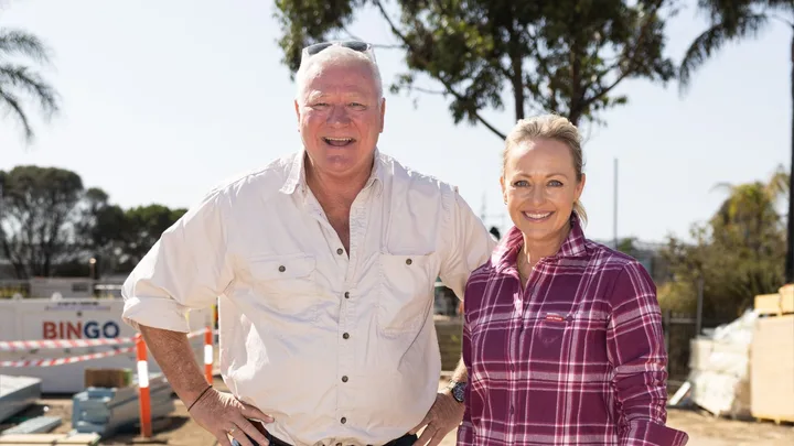 Mature man and woman smiling at a construction site, he wears a light shirt, she wears a plaid shirt; trees in background.