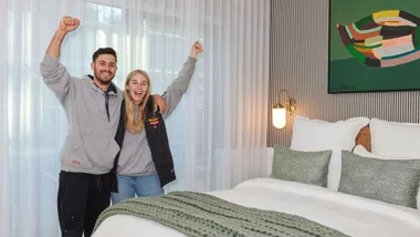 A joyful couple in casual wear celebrates in a modern, well-decorated bedroom with artwork and soft lighting.