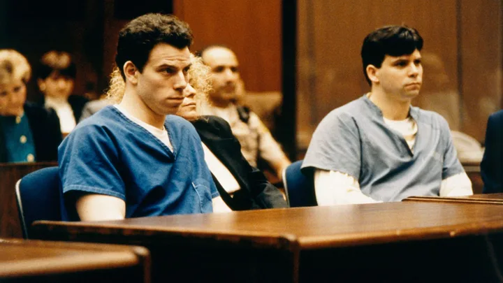 Two men in blue prison uniforms sit in a courtroom with serious expressions.
