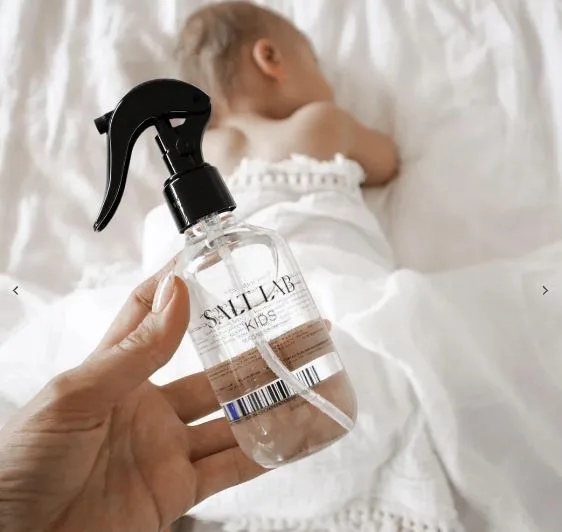 Hand holding a "SALT LAB KIDS" spray bottle with sleeping baby in the background on a white bed.